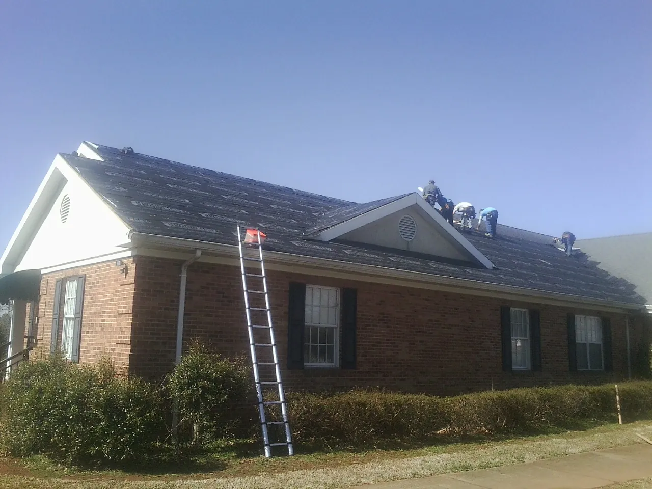 Crew tearing shingles off a brick ranch roof with a ladder set — Metal Roof Repair work by our crew in Loughman