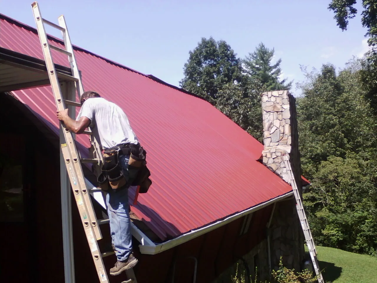 Roofer on a ladder beside a red metal roof and stone chimney — Soffit & Fascia Repair by Roofing Brothers in Loughman